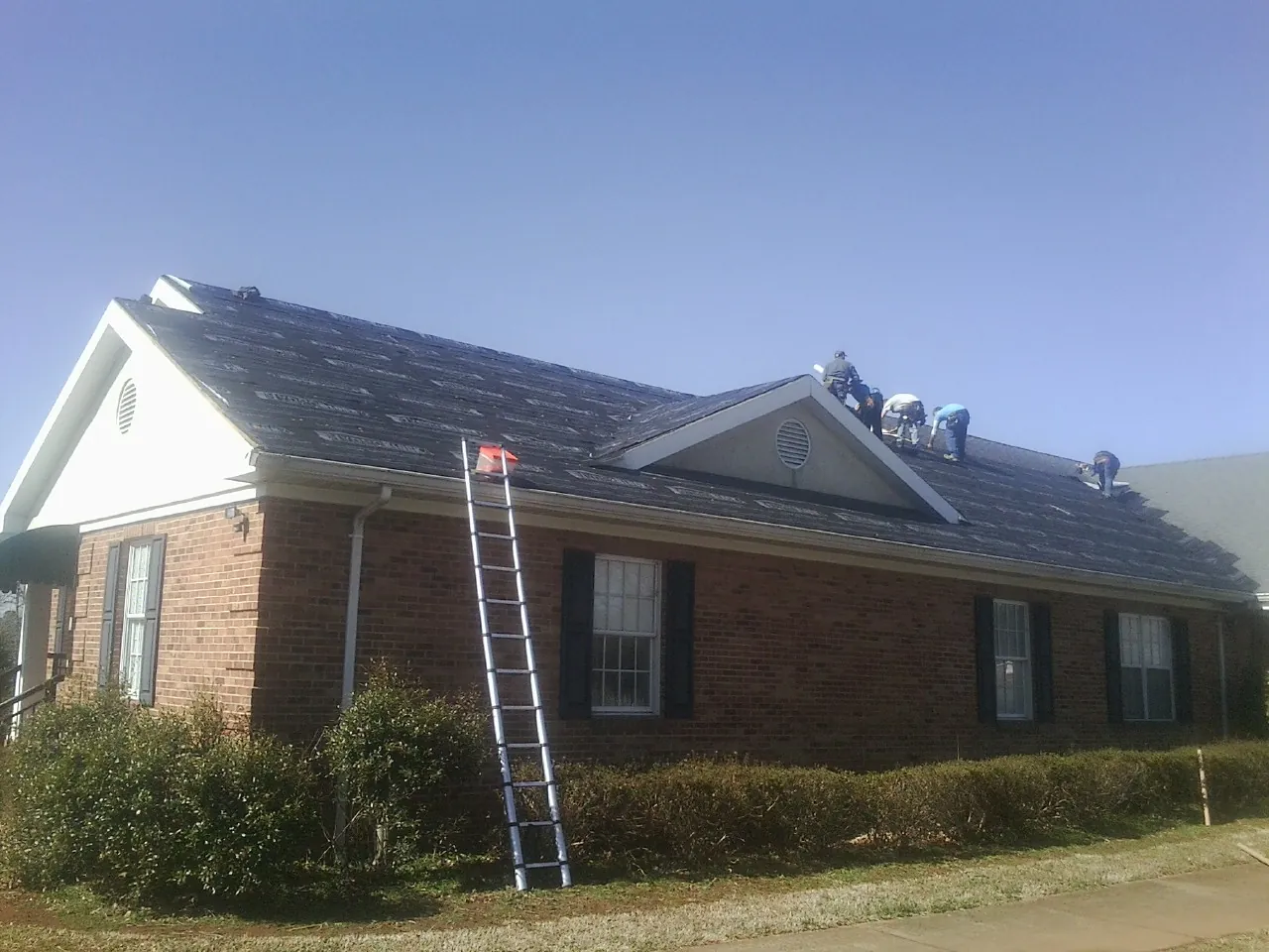 Crew tearing shingles off a brick ranch roof with a ladder set — Emergency Roof Repair work by our crew in Glen Alpine