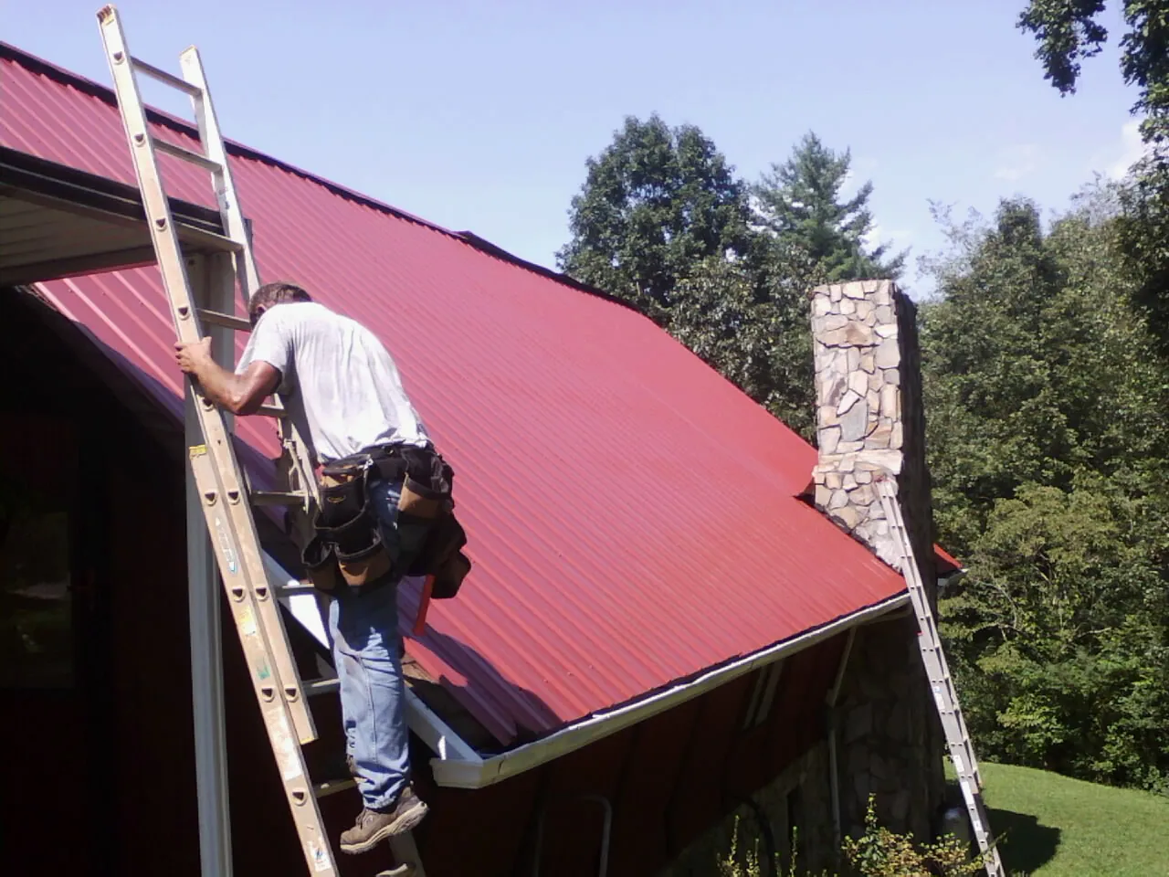 Roofer on a ladder beside a red metal roof and stone chimney — Soffit & Fascia Repair by Roofing Brothers in Glen Alpine